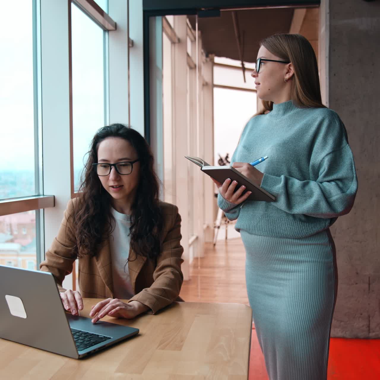 Conversation between the female colleagues. Brunette sits at laptop and blonde lady takes notes into paper notebook