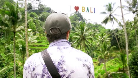 Man-in-cap-standing-in-the-Tegallalang-Rice-Terrace,-"I-heart-Bali"-sign-overhead