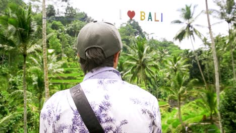 hombre con gorra parado en la terraza de arroz de tegallalang, cartel de &quot;i heart bali&quot; en lo alto