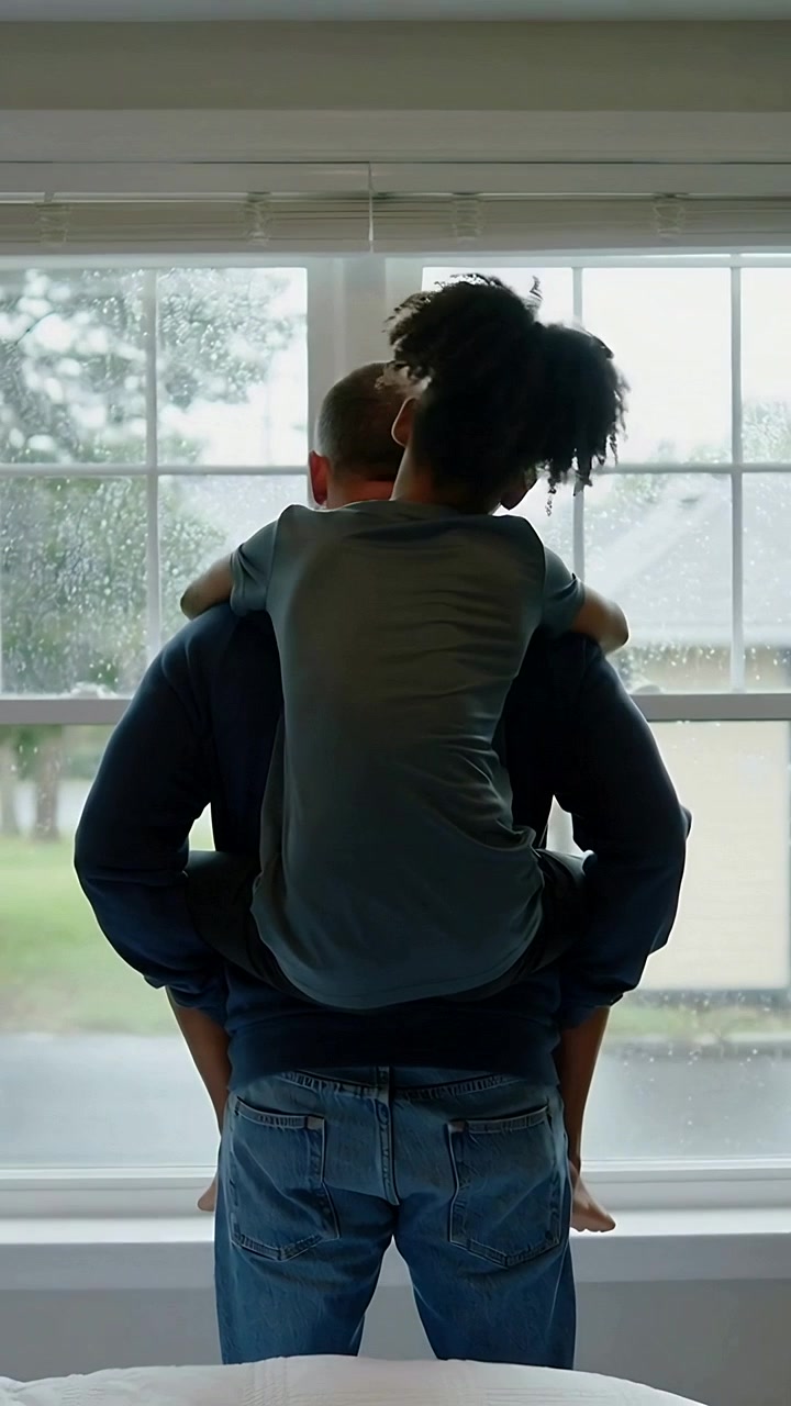 Vertical video: Watching rain falling, hooded dad holding child on shoulders at home window, drops