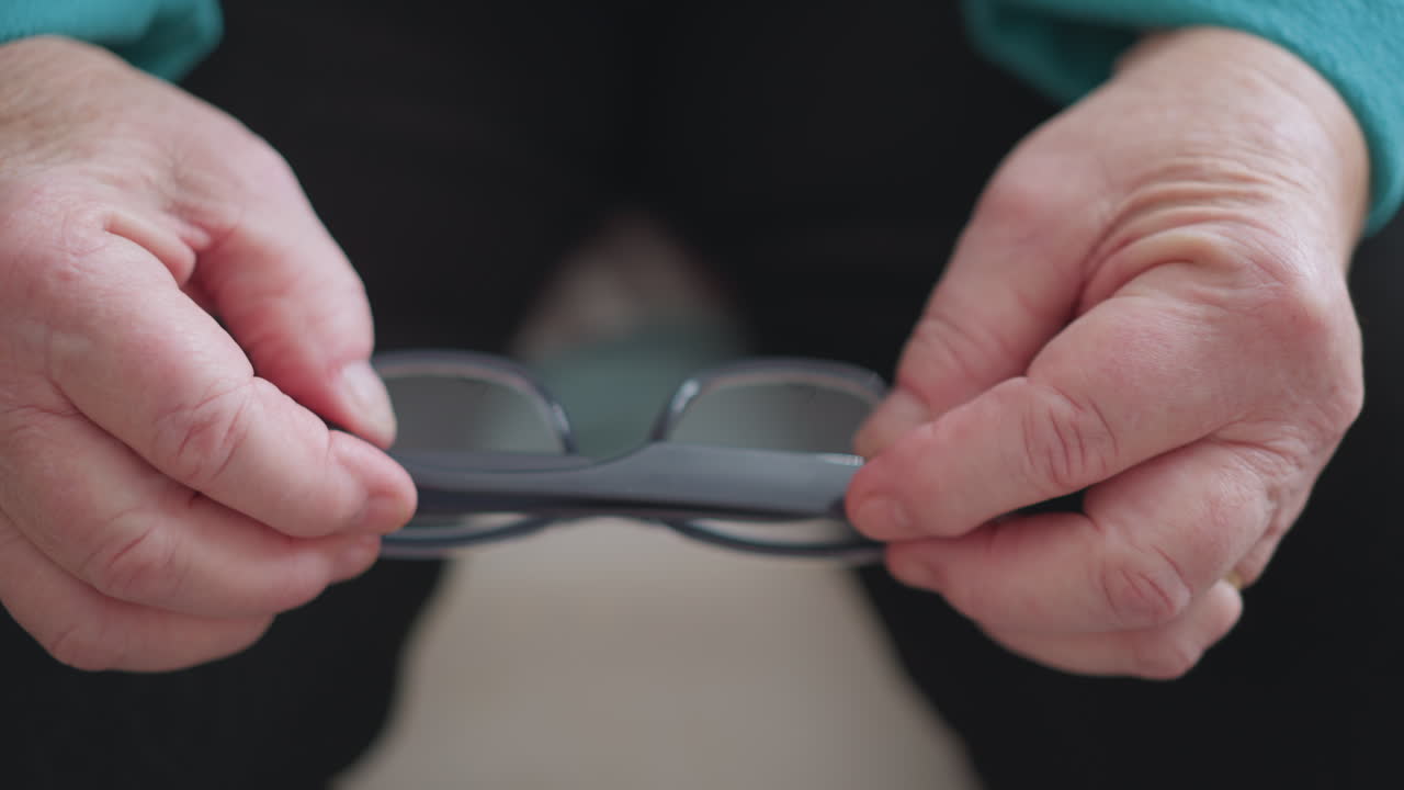 Lonely pensioner in green teal sweater adjusting glasses with both hands while sitting. Close-up of hands gripping eyeglasses, showing an elderly person s focus and care in daily routine
