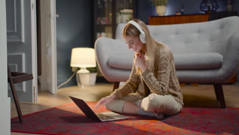 happy young woman with wireless headphones chatting on laptop computer while sitting on floor at home