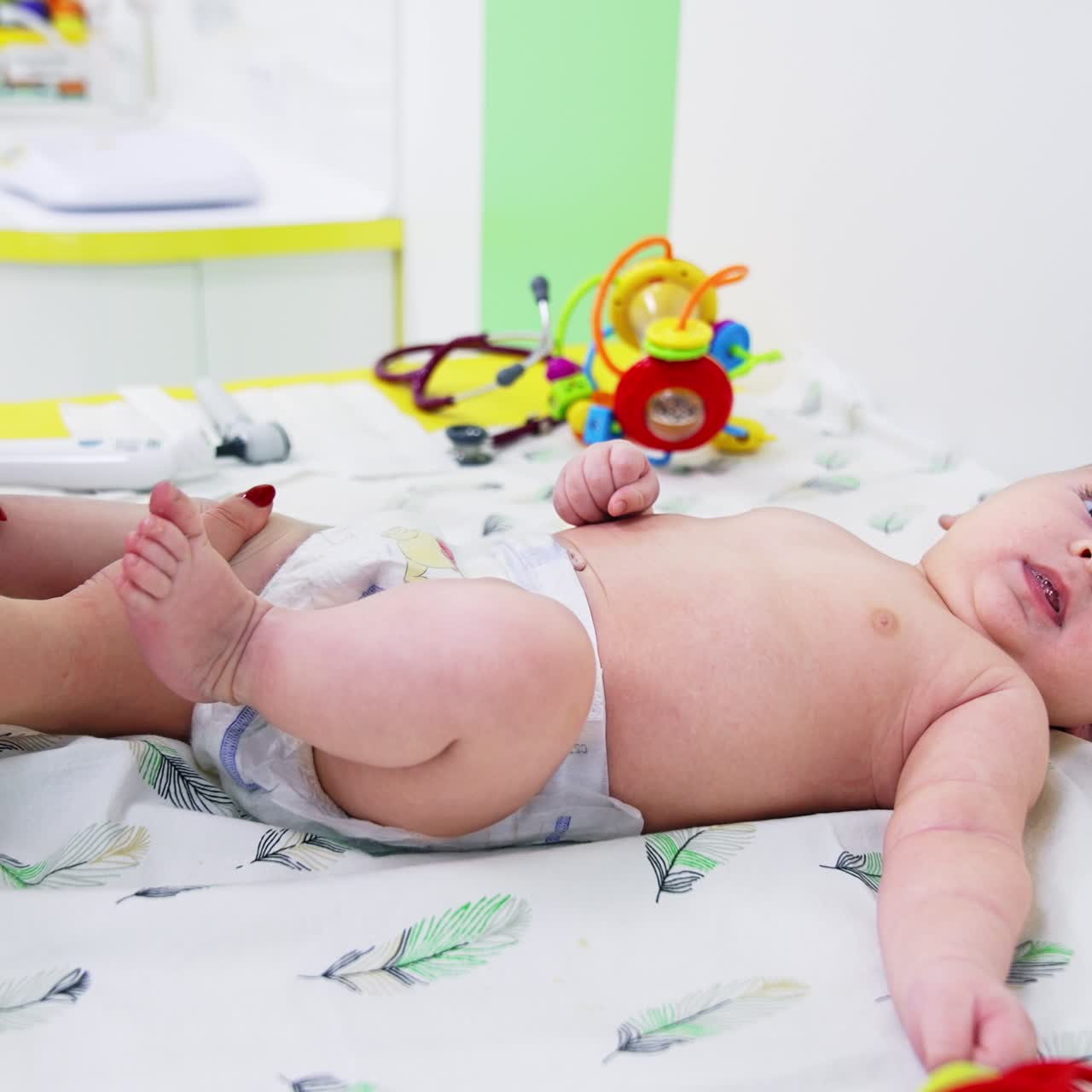 Erkek bebek cerrah kontrolünde. Kadın doktor bebeğin bacaklarını ve dizlerini muayene ediyor. Arka planda oyuncaklar.