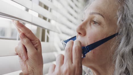 Senior-caucasian-woman-taking-off-glasses-and-looking-through-blinds-out-of-window,-slow-motion