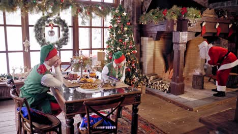 santa claus and two elves are preparing tea. santa claus with boiling teapot, curious boy and girl wear elves clothes. happy elves in fairy costumes sitting near the table. santa near christmas fireplace.