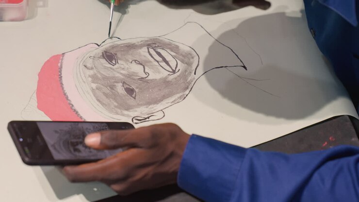 Artista che si riferisce a un ritratto dipinto nero con il telefono, una mano che tiene lo smartphone sopra la carta mentre la tecnica a puntini con inchiostro crea un viso sorridente con un cappello rosso. Movimento puntinato costante, su un piano da studio, contemporaneo.
