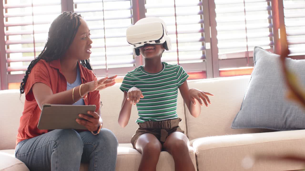 African woman and child sitting on sofa at home with tablet tapping, guiding VR headset play
