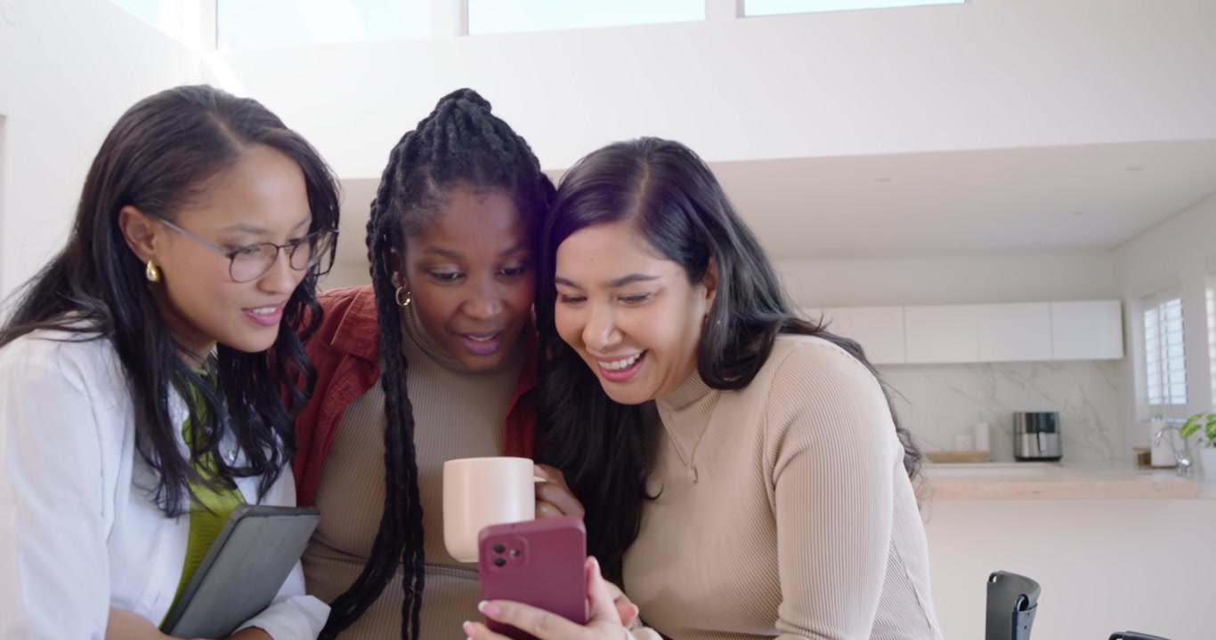Diverse women team leaning laughing in kitchen as right woman tapping pink phone sharing post