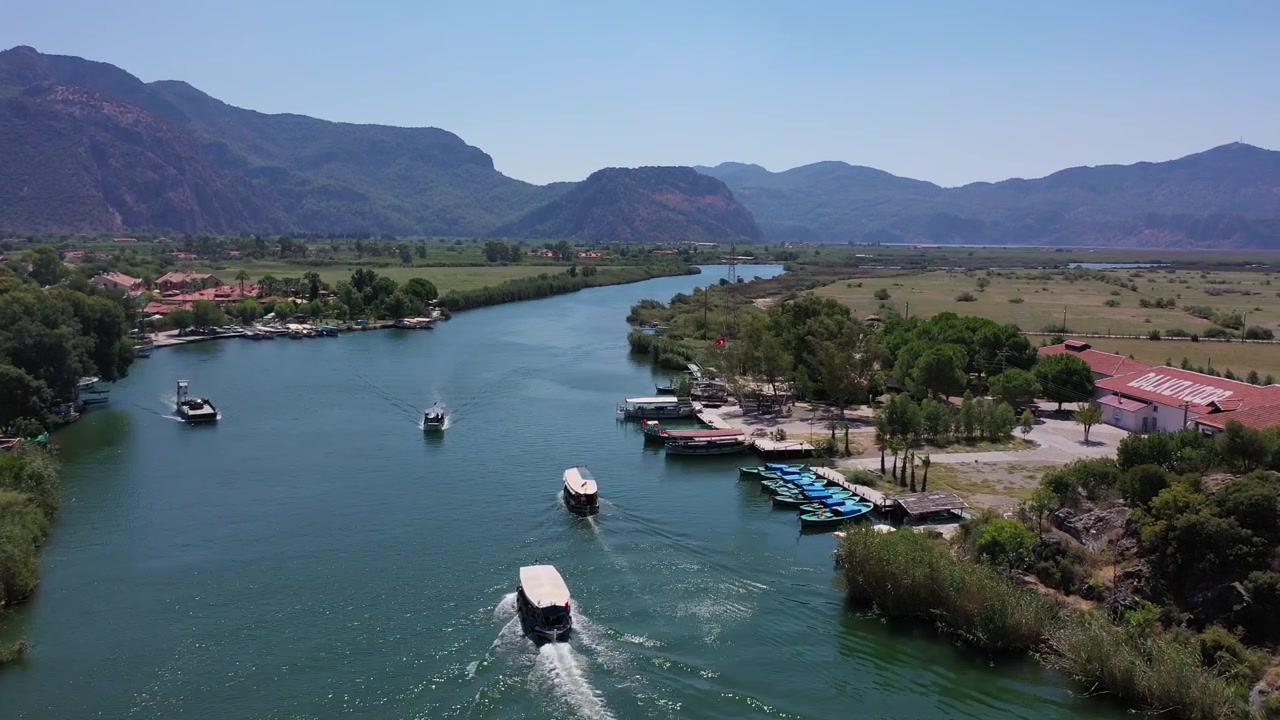 Parlak güneşli bir günde, Türkiye'nin Muğla ilindeki uzak dağlara doğru, ahşap iskelelerin ve sazlıkların yanından, yeşil Dalyan Nehri deltasında seyreden tur teknelerinin manzaralı yüksek havadan drone görünümü.
