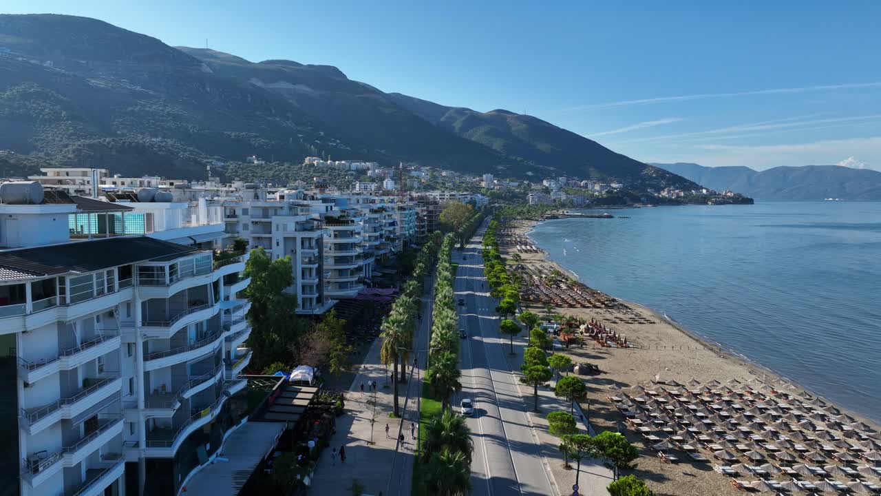 Coastal promenade and beach in Vlore, Albania with mountain views and sunny skies, aerial dolly at early morning past apartments along palm tree road