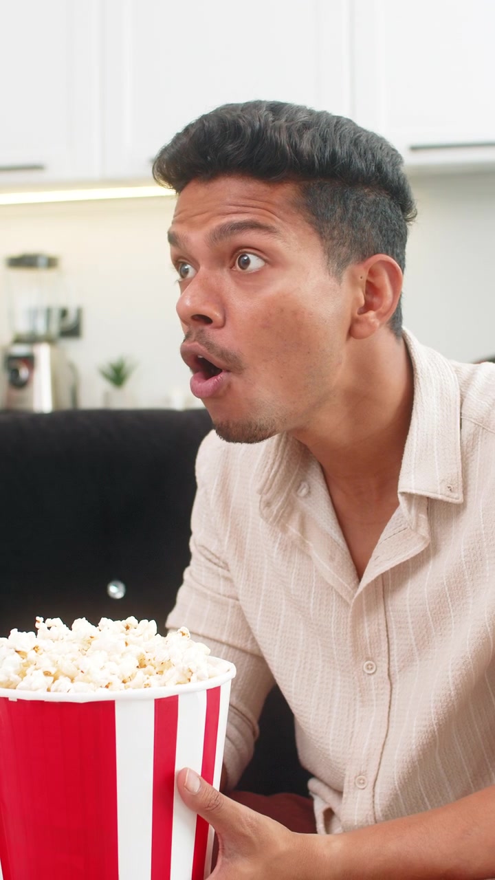 Uomo indiano guarda il trailer di un film in TV, mangia popcorn, ride e reagisce a momenti divertenti, commentando ad alta voce.