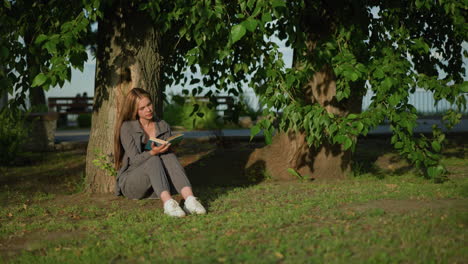 lady sitting outdoor head slightly tilted leaning against tree on grassy field, reading book thoughtfully, tree leaves sway gently in breeze background features greenery and slightly blurred iron rail