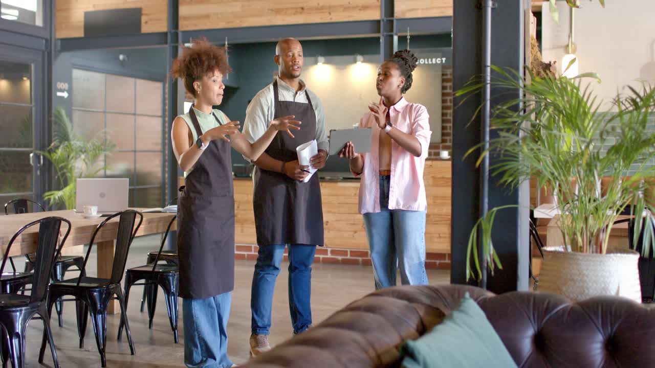Equipe afro-americana de cafeteria planejando, gesticulando para a esquerda, apontando para um tablet ou papéis, dois usando aventais pretos