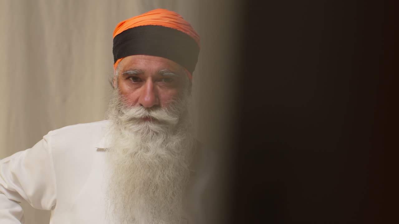 fotografía de estudio de un anciano sikh con barba usando la aguja salai al poner el cabello en el turbante al mirar en el espejo contra un fondo plano