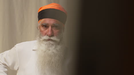 studio-aufnahme eines älteren sikh-mannes mit bart, der eine salai-nadel benutzt, wenn er seine haare in einen turban steckt, während er im spiegel gegen einen einfachen hintergrund schaut