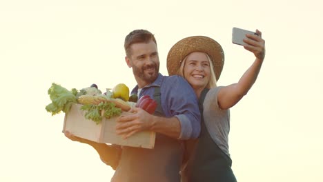 Un-Par-De-Jóvenes-Granjeros-Guapos-Y-Felices-Sonriendo-Y-Posando-Para-La-Cámara-Del-Smartphone-Mientras-Toman-Una-Foto-Selfie-Con-Verduras-Cosechadas-En-Las-Manos