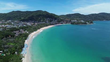 drone zooms in over curving coastline with turquoise waters, beach, forest and town in bright daylight