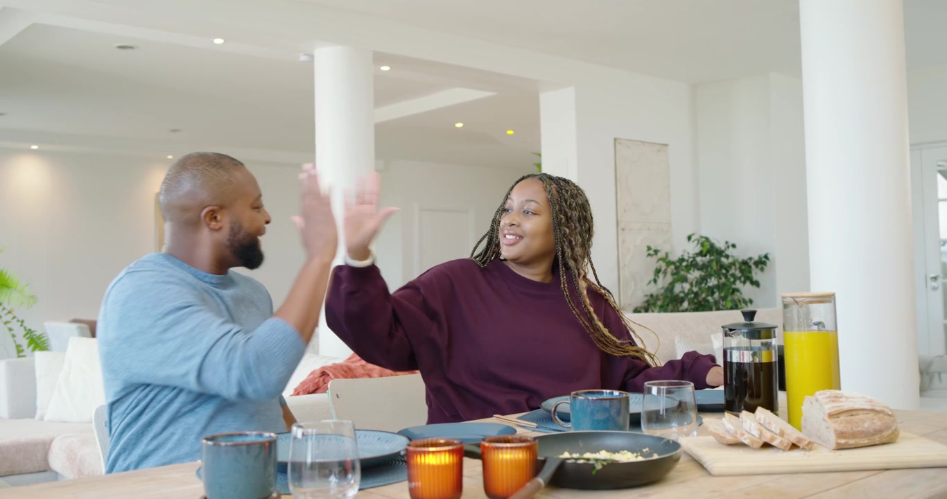 African couple carrying frying pan and board of bagels, plating eggs at table preparing breakfast