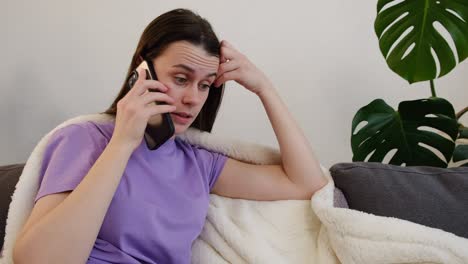 close up of upset sad young caucasian woman speaks on smartphone sitting on sofa in living room at home, gets bad news. adult tired millennial lady covered plaid talking on phone sit on grey couch