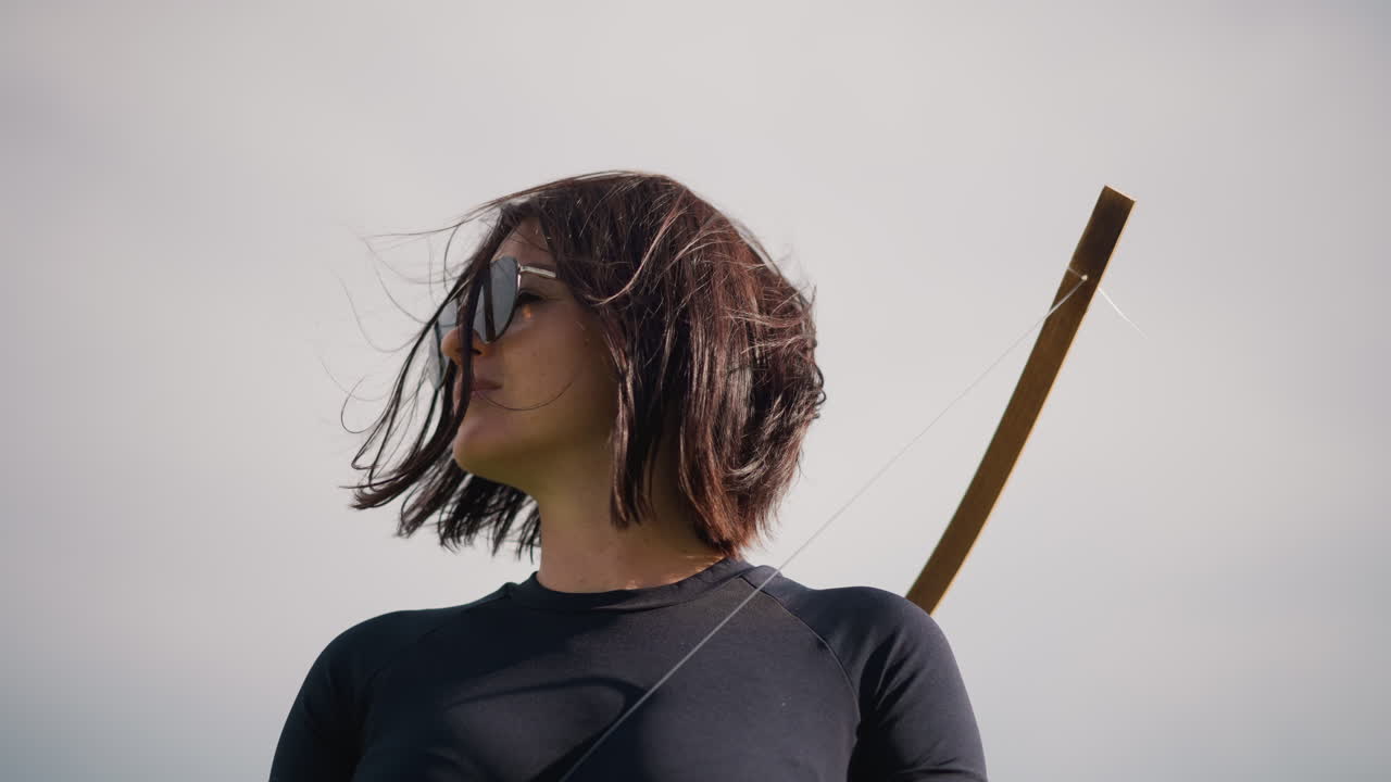 Donna con arco sotto il cielo aperto, capelli al vento e occhiali da sole, ritratto cinematografico ad angolo basso con cinturino in pelle e bastone di legno visibile, nuvole suggestive, sguardo determinato, postura potente, atmosfera