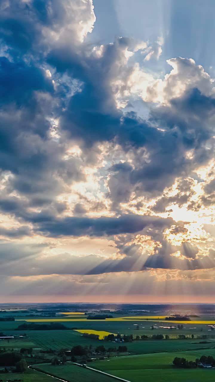 Beautiful aerial view captures dramatic, textured clouds with heavenly sunbeams breaking through, illuminating vast yellow canola fields and green farmland in the picturesque countryside - timelapse