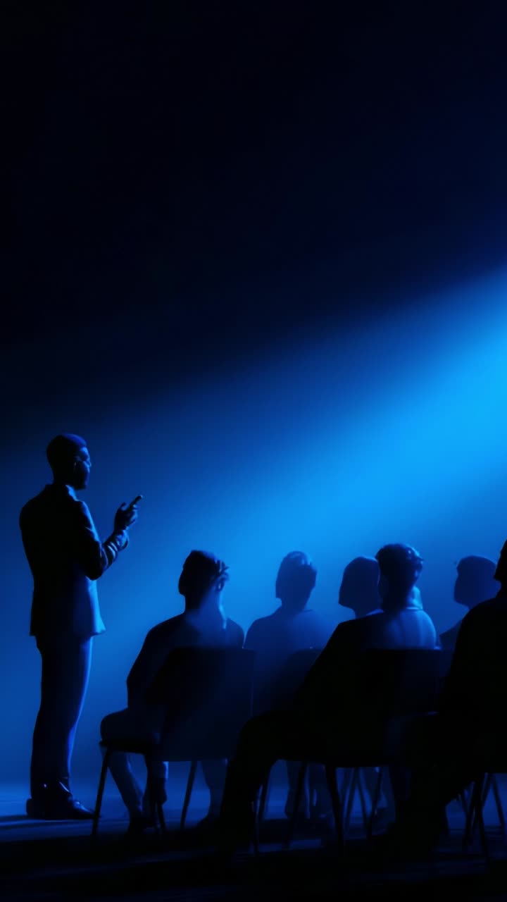 A speaker captivates an audience under dramatic blue lighting, creating an atmosphere of engagement and anticipation in an intimate setting with shadowy figures