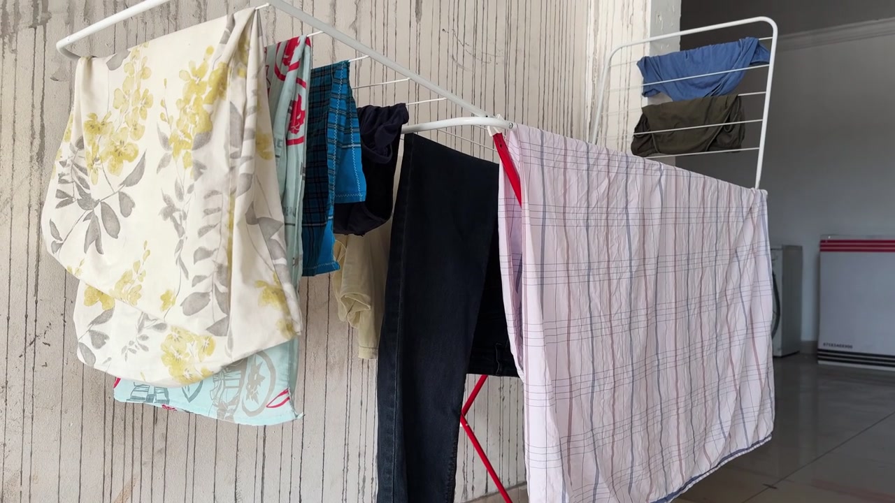 Washed household laundry hangs drying on a white metal rack in a domestic indoor utility space. Natural daylight illuminates the patterned fabrics, shirts, and dark pants in this everyday chore scene.