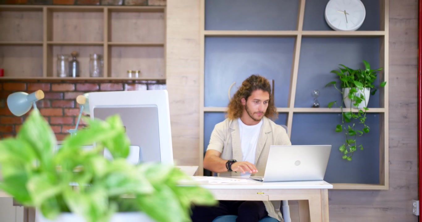 Uomo adulto sui vent'anni che si prepara per il lavoro, in piedi alla scrivania, prende la sedia, si siede e digita sul laptop