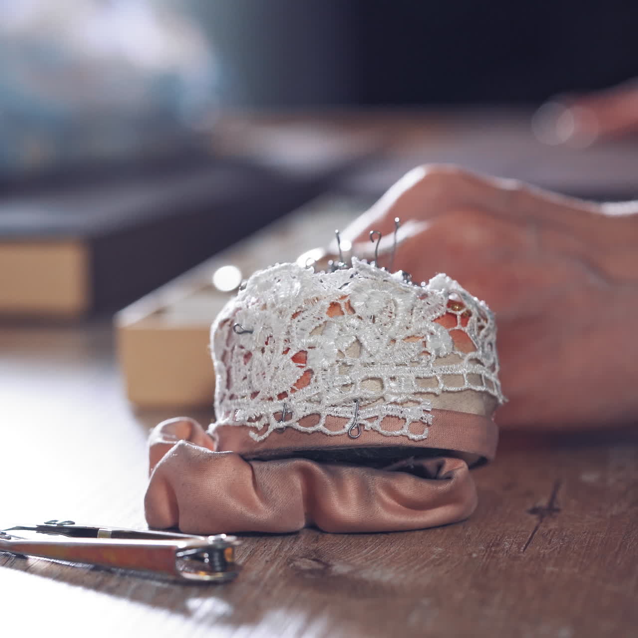 Sewing pins in a white lace sponge for dressmaker on the wooden table. Nice pincushion on the table on the background of tailor hands working in workshop. Close-up.