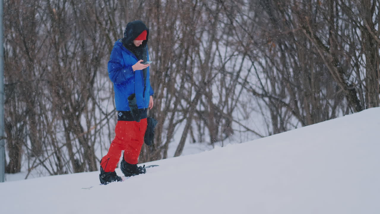 un snowboarder masculino monta la tabla en el esquí hasta la pista de nieve y escribe mensajes en el teléfono inteligente a sus amigos.