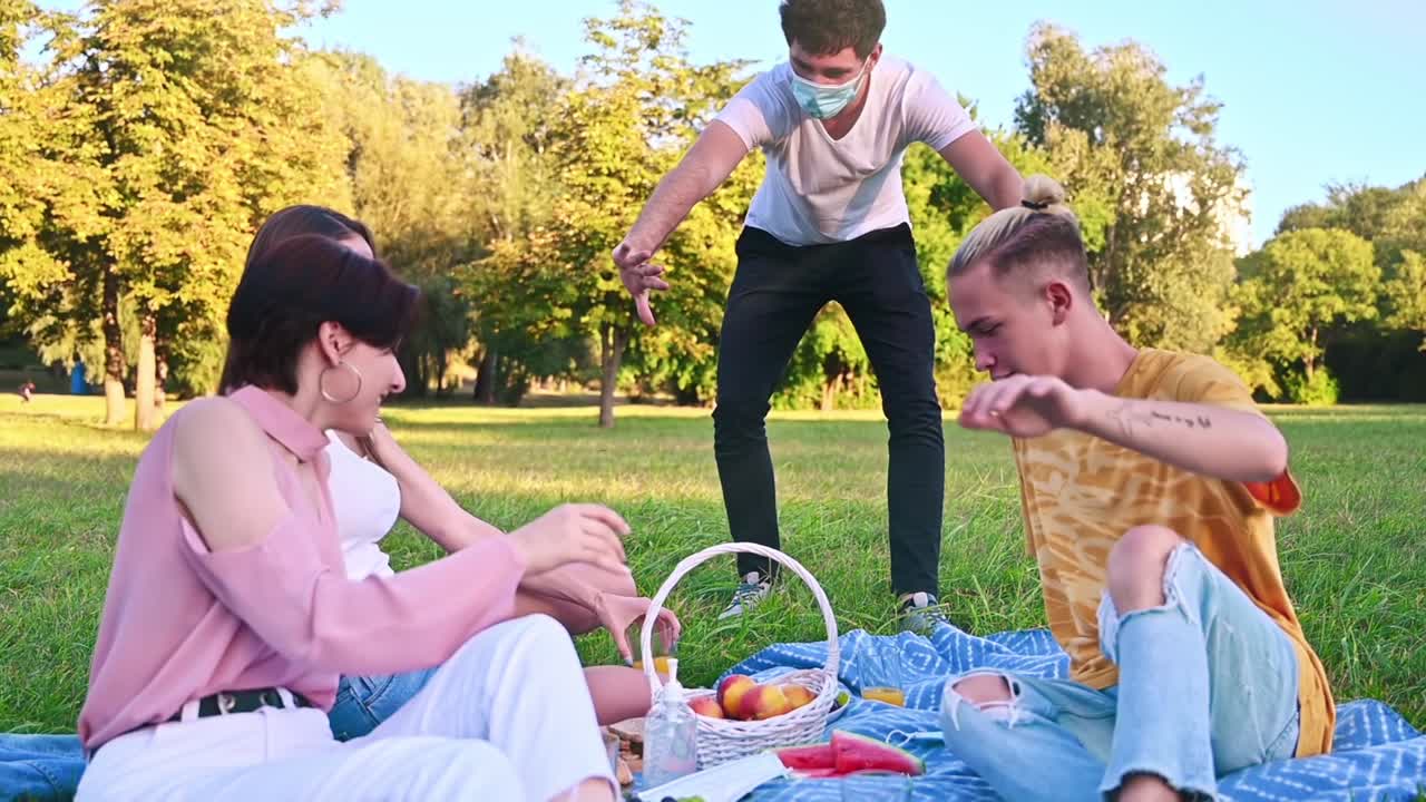 Grupo de amigos divirtiéndose en una manta de picnic con comida en un parque durante la pandemia. Un joven se acerca y les dice a sus amigos que mantengan la distancia social. Idea sobre el coronavirus. Cámara lenta