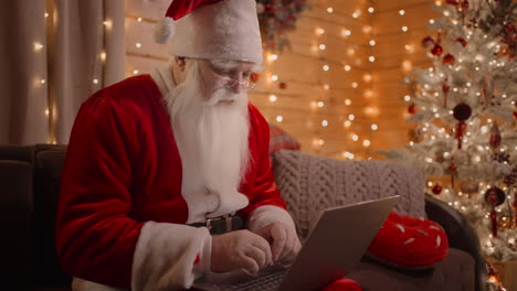 a portrait of a real santa claus working on a laptop walking on the couch at home on christmas eve. santa responds to emails browses the internet bank and works on a laptop