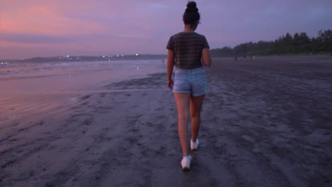 Vista-Panorámica-De-Una-Mujer-Joven-Caminando-En-Una-Playa-De-Arena-Junto-Al-Océano,-Durante-Una-Puesta-De-Sol-Púrpura