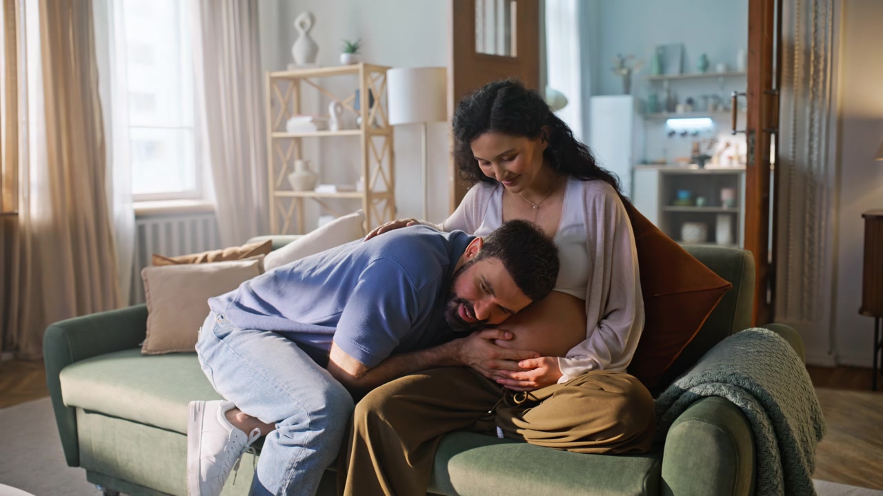 Future father listening pregnant belly home. Couple bonding smiling on sofa.
