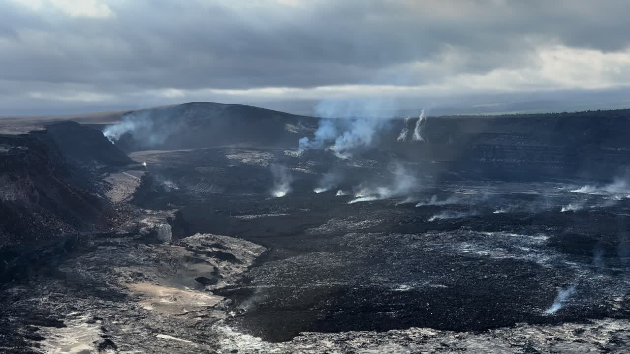Vista statica 4K del cratere Kīlauea che mostra numerosi sfoghi di vapore che si innalzano da campi di lava scuri, rivelando un'ampia attività vulcanica geotermica.