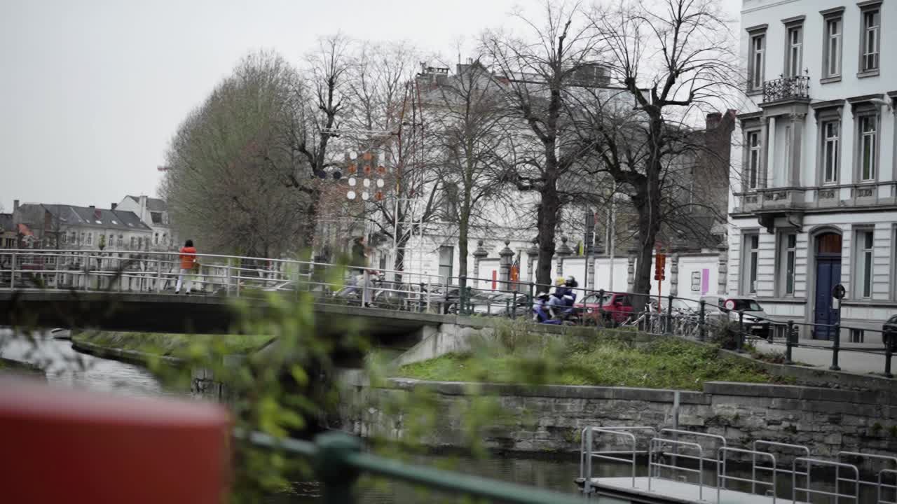 Guy with passenger rides a scooter on a bridge in beautiful city Ghent, Belgium. Helmet protection. Other passengers passing by.