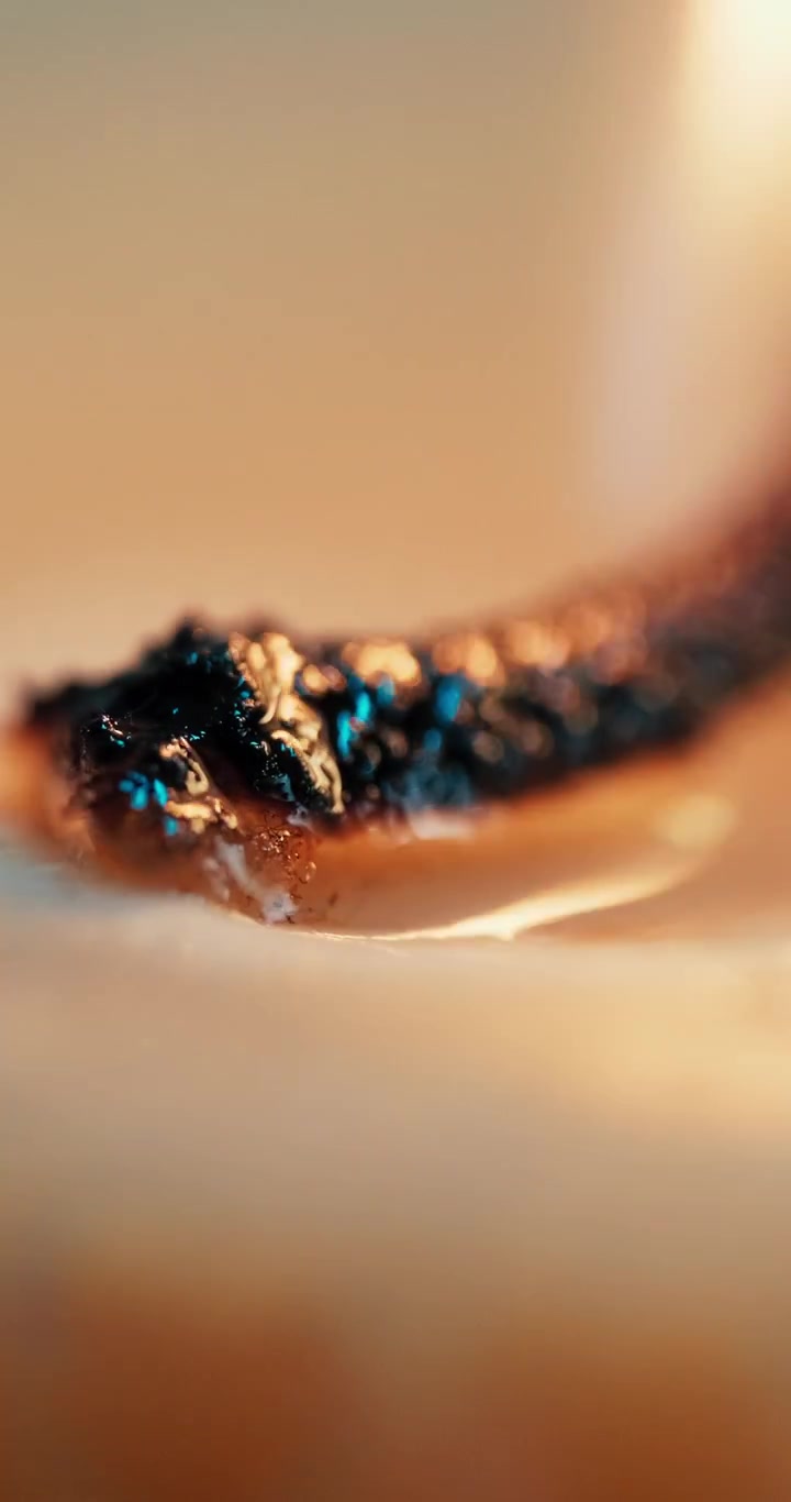 Una ripresa macro cinematografica di uno stoppino di candela carbonizzato che brucia nella cera fusa, con illuminazione calda dell'ora d'oro e un'estetica minimalista per contenuti di benessere e consapevolezza.