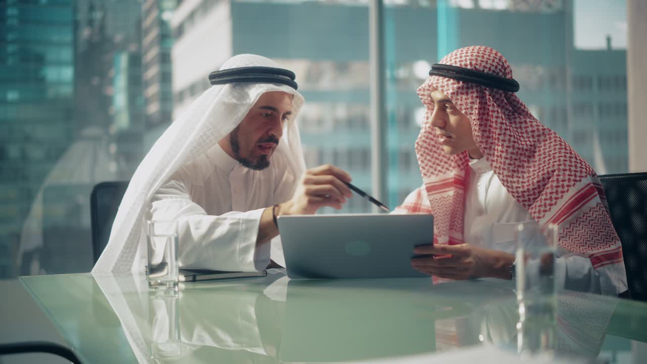 dos empresarios emiratíes exitosos con trajes tradicionales blancos sentados en una reunión de oficina, negociando y hablando sobre oportunidades financieras, usando una computadora portátil.