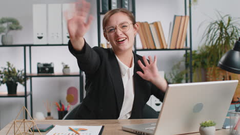 businesswoman working on laptop smiling friendly at camera and waving hand gesturing hello at office