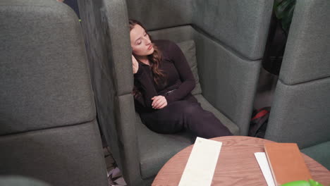 relaxed young woman in black top leans against cushioned booth, holding smartphone to her ear while listening attentively. cozy lounge ambiance features brown round table, visible plant and brick wall