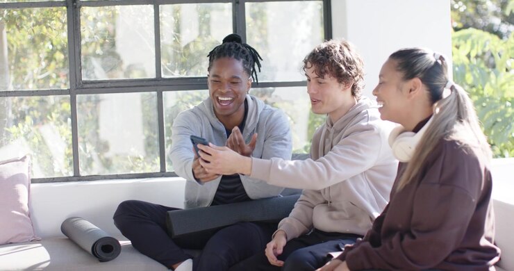Diverse friends leaning in while left man holding smartphone and sharing funny clip on sunlit bench