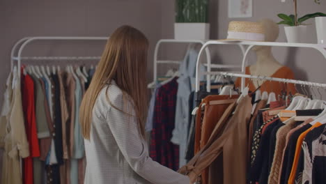 joven hermosa en una tienda de ropa buscando vestidos y suéteres en el mostrador. para tomar de una percha y para poner a sí misma sonriendo.