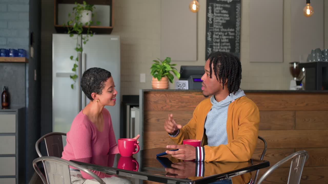 After barista exiting, couple checking smartphone, lifting red mugs, leaning in conversing at cafe