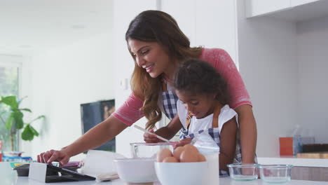 hispanic mother and daughter in kitchen following cake recipe on digital tablet together
