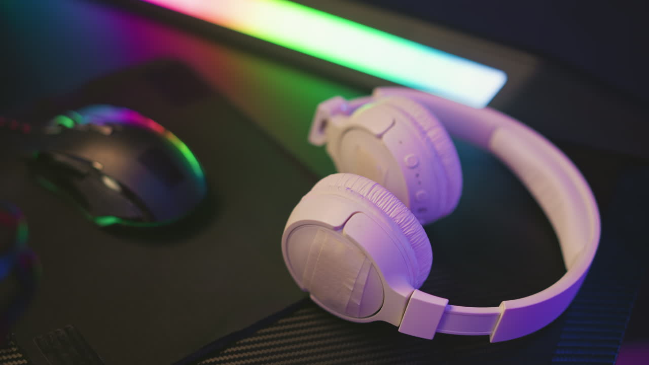 Close up white headset and black mouse placed on dark surface near bright multicolor LED light bar with vibrant reflections highlighting modern desktop setup in colorful
