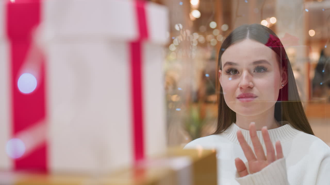 Close-up of beautifully wrapped gift box displayed in store window as lady admires it through glass with people walking by in busy shopping mall