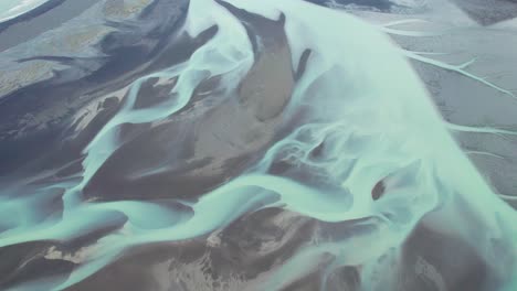 Aerial-view-of-the-colorful-blue-glacier-rivers-from-above-in-Iceland-during-summer,-showcasing-intricate-patterns-and-vibrant-hues-against-the-black-volcanic-sands