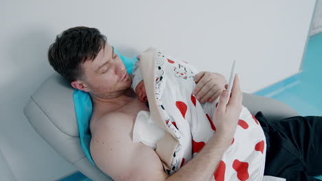 young caucasian father holding his newborn son on the chest. happy dad takes the phone and looks at the screen.