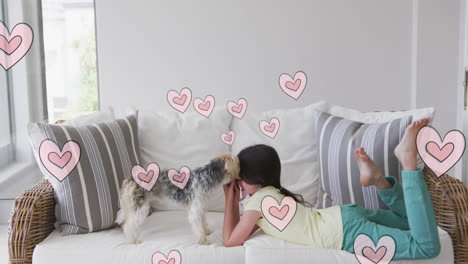 animation of hearts moving over caucasian girl wearing face mask playing with her pet dog