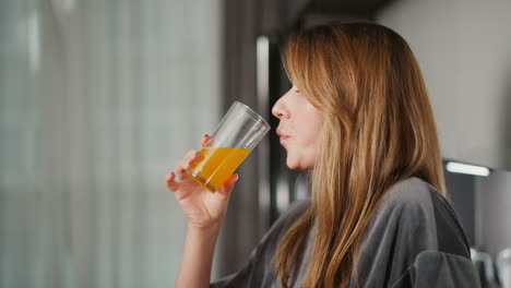 side view of lady with long hair drinking orange juice from transparent glass in bright kitchen, natural daylight softly illuminates scene, with modern furnishings